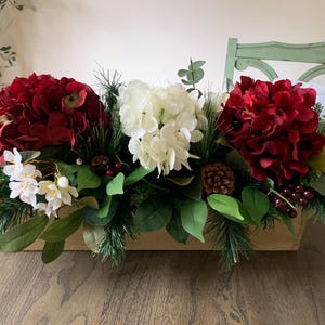 May include: A wooden box centerpiece with red and white hydrangea flowers, pine sprigs, pine cones, and berries.