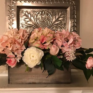 May include: A wooden box overflowing with pink and white faux flowers, including hydrangeas, roses, and ranunculus. The arrangement is set against a decorative silver metal wall hanging with an embossed design.