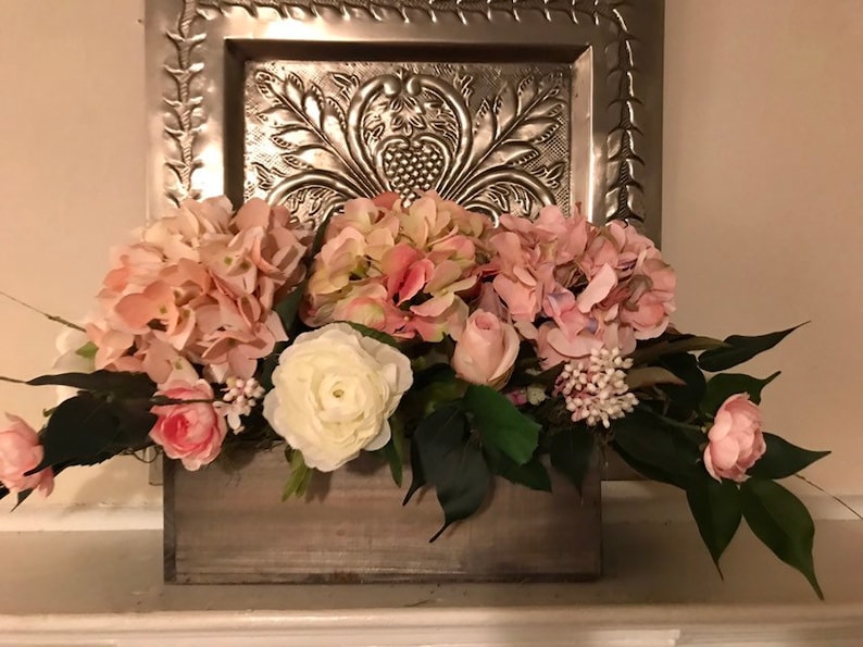 May include: A wooden box overflowing with pink and white faux flowers, including hydrangeas, roses, and ranunculus. The arrangement is set against a decorative silver metal wall hanging with an embossed design.