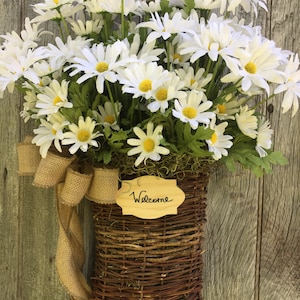 May include: A brown wicker basket filled with white daisies and a burlap bow. A wooden sign with the word "Welcome" is attached to the basket.