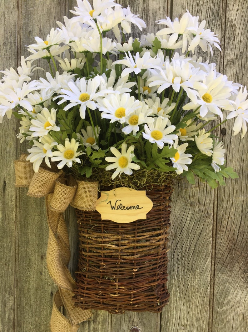 May include: A brown wicker basket filled with white daisies and a burlap bow. A wooden sign with the word "Welcome" is attached to the basket.