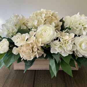 May include: A rectangular wooden box overflowing with a lush arrangement of white and cream-colored flowers. The arrangement includes hydrangeas, roses, and greenery, creating a full and elegant display. Perfect for home decor.