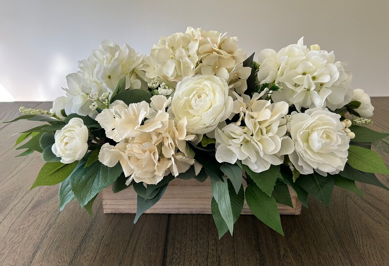 May include: A rectangular wooden box overflowing with a lush arrangement of white and cream-colored flowers. The arrangement includes hydrangeas, roses, and greenery, creating a full and elegant display. Perfect for home decor.