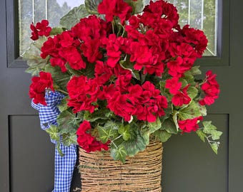 May include: A rectangular woven twig basket filled with vibrant red geranium flowers and green leaves. A blue and white checkered gingham ribbon is tied to the left side of the basket, with two long fabric tails hanging down from the arrangement.