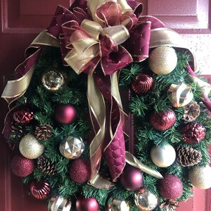 May include: A Christmas wreath with a burgundy and gold bow, decorated with burgundy and gold ornaments, pine cones, and sprigs of greenery.