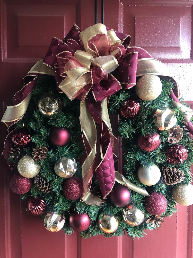 May include: A Christmas wreath with a burgundy and gold bow, decorated with burgundy and gold ornaments, pine cones, and sprigs of greenery.