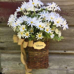 May include: A wicker basket filled with white daisies and a burlap bow, hanging on a wooden door with a sign that says "Welcome".