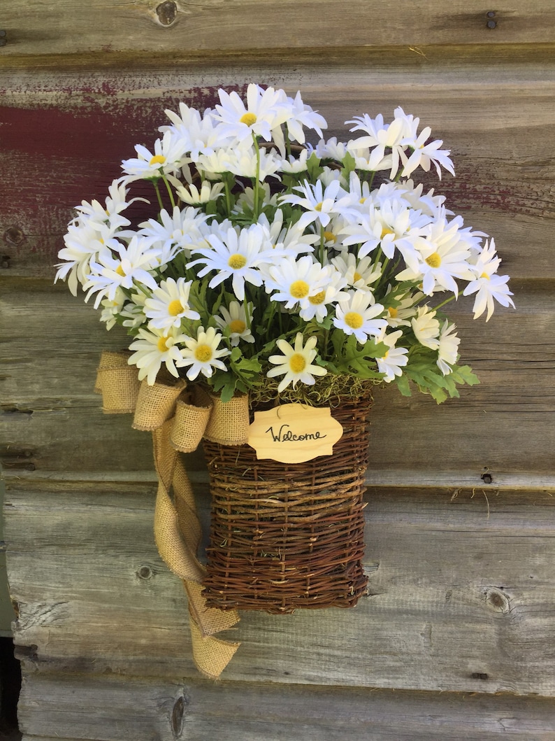 May include: A wicker basket filled with white daisies and a burlap bow, hanging on a wooden door with a sign that says "Welcome".
