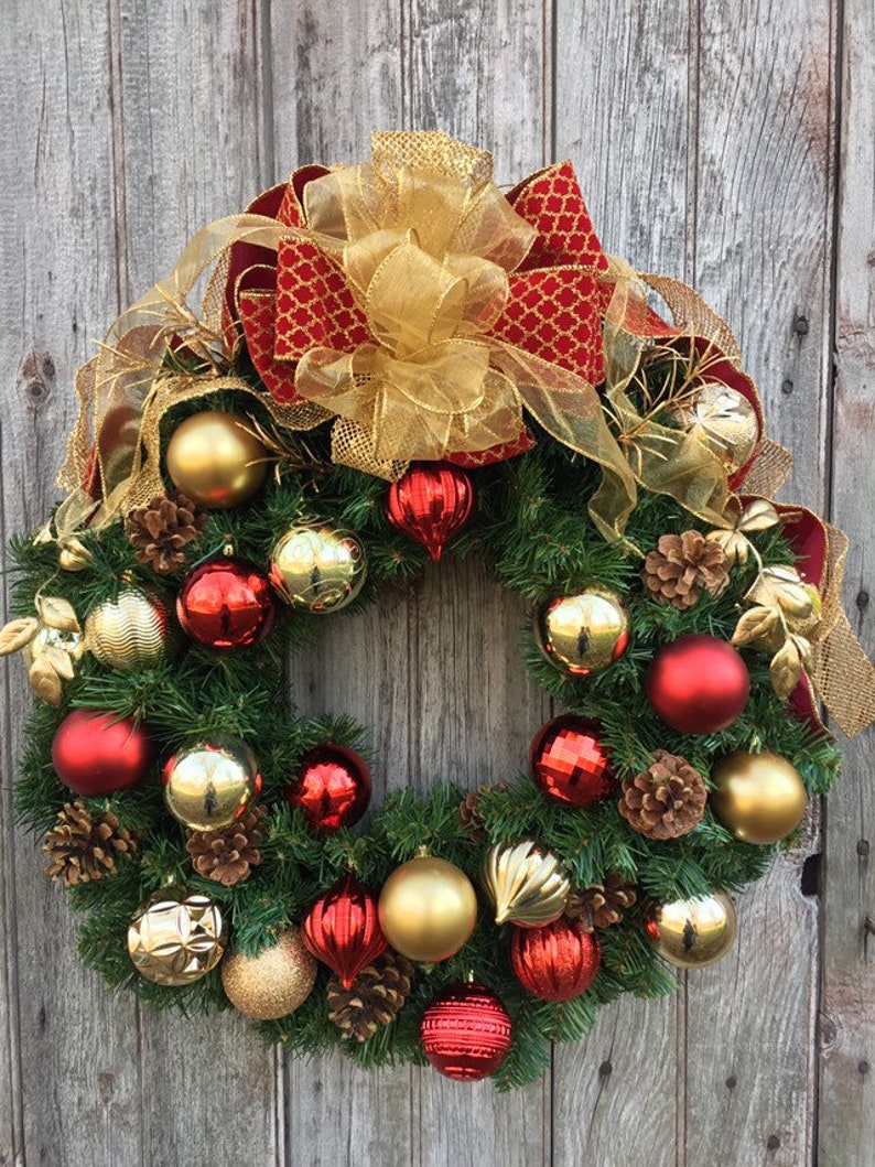 May include: A Christmas wreath decorated with red and gold ornaments, pine cones, and a large gold and red bow.