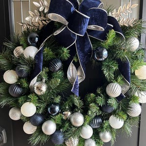 May include: A Christmas wreath with a navy blue velvet bow and silver trim. The wreath is decorated with silver, white, and dark blue ornaments.