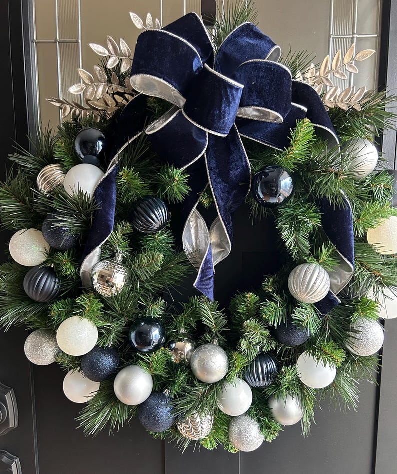 May include: A Christmas wreath with a navy blue velvet bow and silver trim. The wreath is decorated with silver, white, and dark blue ornaments.