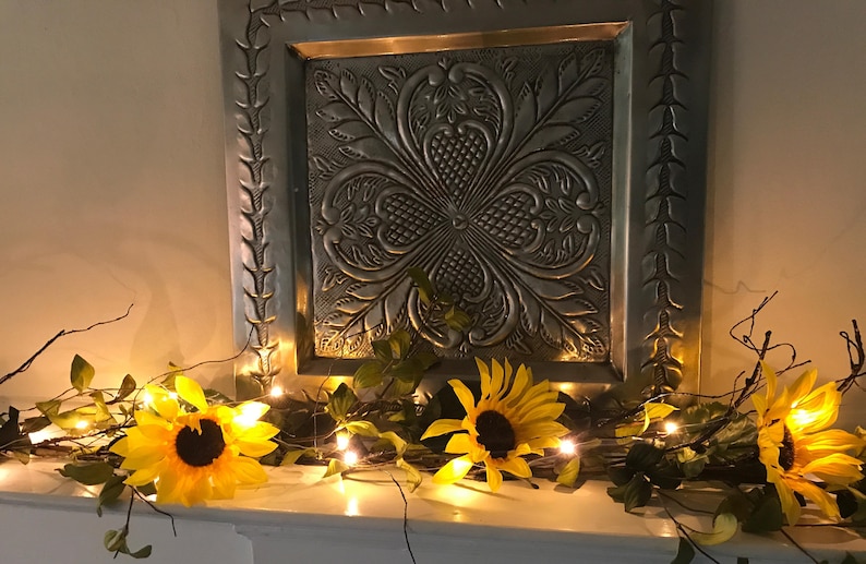 以下が含まれることがあります： 花柄の装飾的な金属製の壁パネルが、フェイクのひまわりと緑の葉のガーランドの上にあり、ストリングライトが飾られています。