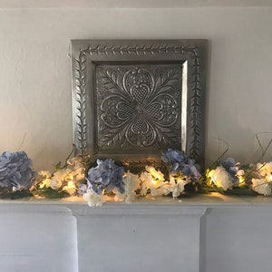 May include: A white fireplace mantel decorated with a garland of blue and white flowers and white fairy lights. A silver metal wall decor with a floral design is above the garland.