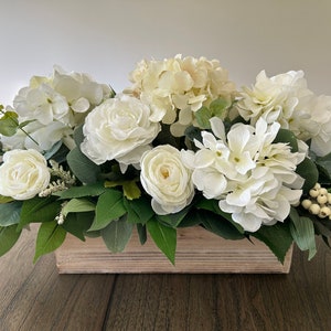 May include: A rectangular wooden box overflowing with white faux flowers, including hydrangeas and roses, accented by green leaves and small white berries. The arrangement is a centerpiece for home decor.