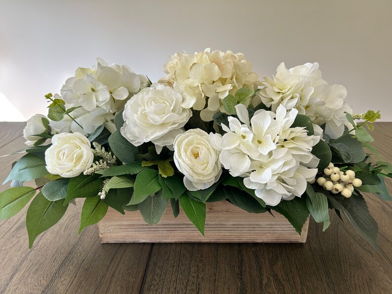 May include: A rectangular wooden box overflowing with white faux flowers, including hydrangeas and roses, accented by green leaves and small white berries. The arrangement is a centerpiece for home decor.