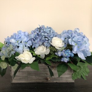 May include: A floral arrangement featuring blue hydrangeas and white roses in a rustic wooden box. Green leaves and foliage provide a natural accent. The arrangement is a centerpiece for home decor.