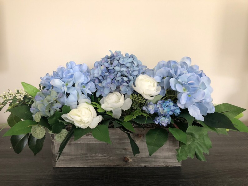 May include: A floral arrangement featuring blue hydrangeas and white roses in a rustic wooden box. Green leaves and foliage provide a natural accent. The arrangement is a centerpiece for home decor.