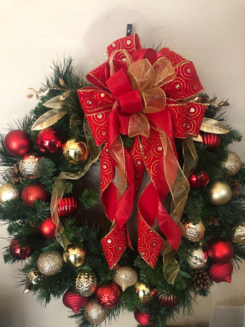 May include: A Christmas wreath decorated with red and gold ornaments, pine branches, and a large red bow with gold accents.