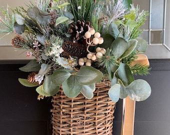 May include: A brown wicker basket filled with a wintery floral arrangement. The arrangement includes pine sprigs, eucalyptus leaves, white berries, and pine cones. A beige ribbon hangs from the basket.
