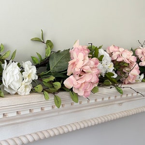 May include: A white mantelpiece decorated with a garland of pink and white artificial flowers. The garland is made of hydrangeas and other small white flowers with green leaves.