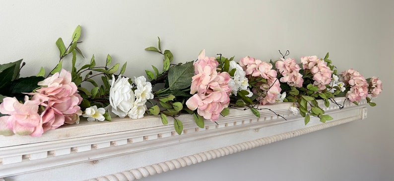 May include: A white mantelpiece decorated with a garland of pink and white artificial flowers. The garland is made of hydrangeas and other small white flowers with green leaves.