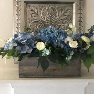 May include: A wooden rectangular planter box filled with blue hydrangeas, white roses, and greenery. The arrangement sits in front of a decorative silver wall hanging. The flowers are a mix of light and dark blue.