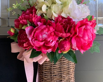 May include: A pink and white floral arrangement in a wicker basket with a pink ribbon. The arrangement includes peonies, hydrangeas, and orchids.