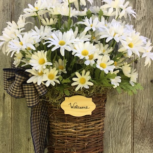 May include: A wicker basket filled with white daisies and a blue and black checkered ribbon. The basket has a wooden sign that says "Welcome".