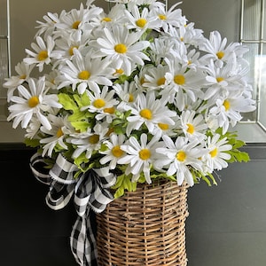 May include: A white daisy wreath with a black and white checkered ribbon bow, hanging in a woven wicker basket.