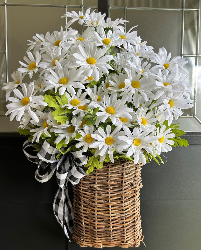 May include: A white daisy wreath with a black and white checkered ribbon bow, hanging in a woven wicker basket.