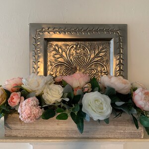 May include: A wooden box centerpiece with a variety of pink and white flowers, including roses and peonies. The box is sitting on a white surface in front of a silver decorative tray.