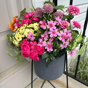 May include: A vibrant floral arrangement in a gray planter on a black metal stand. The arrangement features a variety of colorful flowers, including pink, yellow, and orange blooms, with green foliage. The planter is round and sits on a geometric stand.