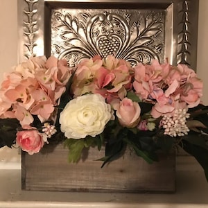 May include: A wooden box overflowing with pink and white faux flowers, including hydrangeas, roses, and other blooms. The arrangement sits in front of a decorative silver metal panel.