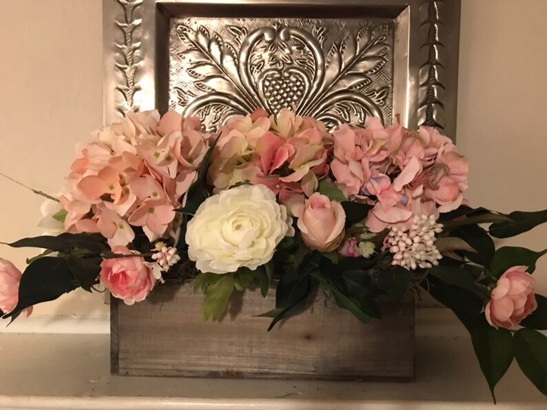 May include: A wooden box overflowing with pink and white faux flowers, including hydrangeas, roses, and other blooms. The arrangement sits in front of a decorative silver metal panel.