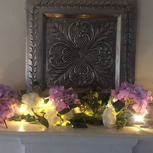 May include: A garland of white and purple flowers with small white lights strung through it. The garland is draped over a decorative silver tray with an embossed design.