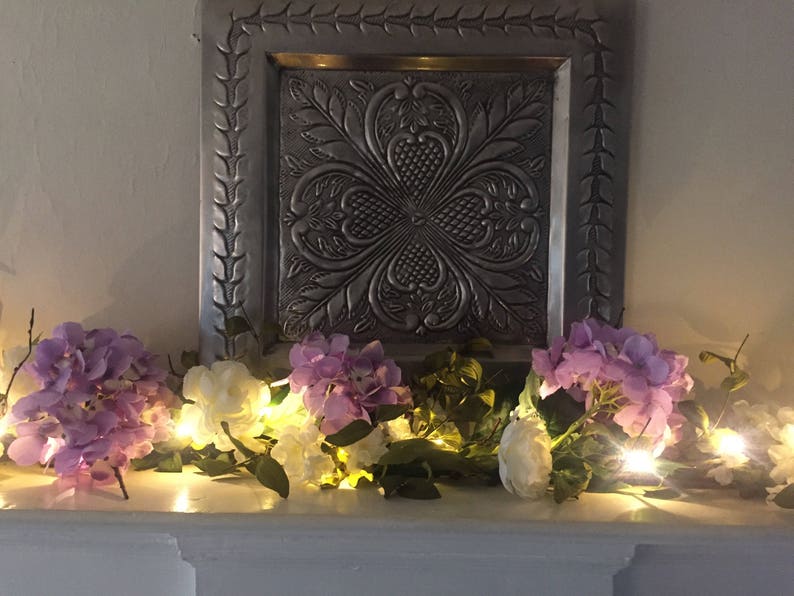 May include: A garland of white and purple flowers with small white lights strung through it. The garland is draped over a decorative silver tray with an embossed design.