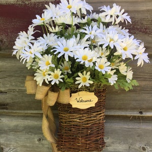 May include: A wicker basket filled with white daisies and a burlap bow hangs on a wooden door. A small wooden sign with the word "Welcome" is attached to the basket.