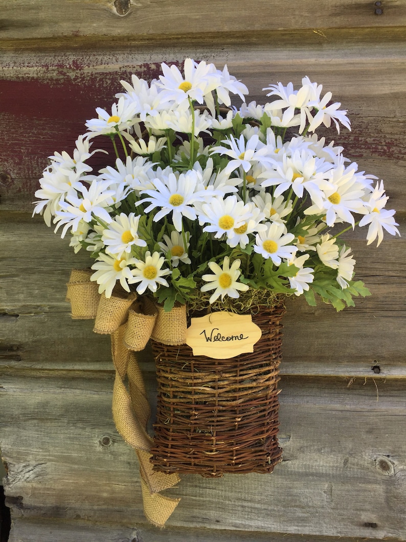 May include: A wicker basket filled with white daisies and a burlap bow hangs on a wooden door. A small wooden sign with the word "Welcome" is attached to the basket.