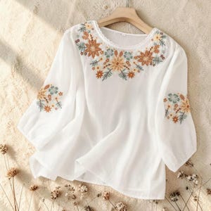 Puede incluir: Una blusa blanca de manga larga con escote redondo y un delicado borde de encaje. La prenda está decorada con intrincados bordados florales en tonos naranja quemado, amarillo mostaza y verde salvia alrededor del cuello y en las mangas.