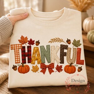 May include: A cream-coloured crewneck sweatshirt featuring the word THANKFUL in large, multi-patterned fabric letters. The design includes orange gingham, red plaid, yellow floral, sage green, quilted cream, and brown polka dot textures with pumpkins, leaves, acorns, and a red bow.