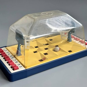 May include: A tabletop basketball game featuring a yellow plastic court with recessed holes, two blue hoops, and a clear plastic dome cover. The base is dark blue with a row of ten red numbered buttons labeled 1 through 0 for gameplay.