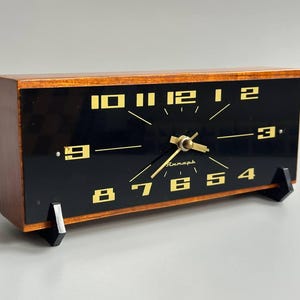 Op de afbeelding: Een rechthoekige tafelklok met een houten frame en een zwarte wijzerplaat. De klok heeft goudkleurige cijfers van 1 tot 12, gouden wijzers en een klein goudkleurig logo. Het wordt ondersteund door twee zwarte kunststof voetjes onderaan.