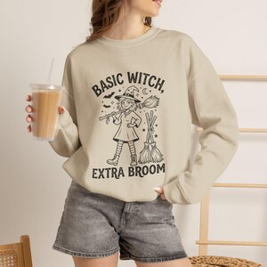 May include: A beige crewneck sweatshirt featuring a black graphic print of a witch wearing a hat and boots, holding a broomstick. The text reads BASIC WITCH, EXTRA BROOM, accompanied by small illustrations of bats, stars, and a crescent moon.