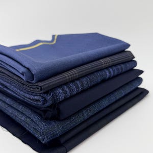 Può includere: Una pila di sei campioni di tessuto piegati in varie tonalità di blu e blu navy. Il pezzo superiore è un blu medio tinta unita con una sottile linea gialla lungo il bordo, seguito da strati di tessuti blu navy a quadri, testurizzati e blu navy.