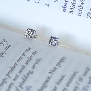Peut inclure: Une paire de petites boucles d'oreilles carrées en argent. L'une présente un motif linéaire texturé, tandis que l'autre affiche un motif d'abeille en relief détaillé. Les boucles d'oreilles sont posées sur les pages d'un livre ouvert.