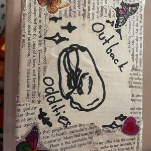 May include: A rectangular box covered in vintage book pages featuring a hand-drawn black ink illustration of a brain. The surface is decorated with colourful butterfly stickers, black star symbols, and a pink heart sticker. The text Outback Oddities is written in black ink.
