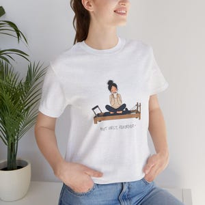 May include: A light grey heather t-shirt featuring a graphic of a person sitting cross-legged on a brown pilates reformer machine while holding a white mug. Below the illustration, the text reads, "BUT FIRST, REFORMER" in black lettering.