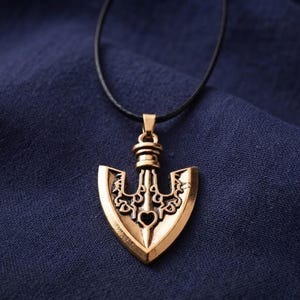 Puede incluir: Un colgante de metal dorado con forma de punta de flecha, con intrincados calados y una pequeña abertura en forma de corazón en el centro. El colgante cuelga de un sencillo cordón negro y presenta un acabado pulido con grabados detallados.
