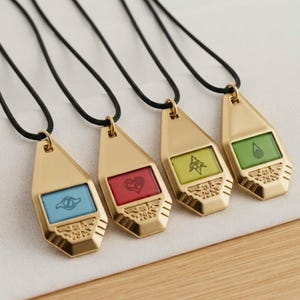 May include: Four gold-toned metal pendants on black cords, each featuring a trapezoidal shape with a central rectangular inlay. The inlays are light blue, red, yellow, and green, each displaying a unique black symbol and a small wing motif.