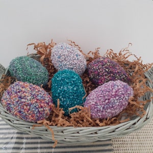 May include: A gray wicker basket filled with colorful, textured, yarn-covered Easter eggs. The eggs are in shades of green, blue, purple, and pink, nestled on brown paper filler. The basket has a curved handle.